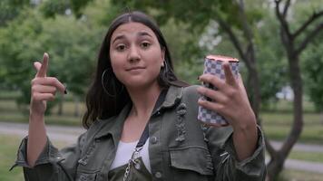 Young hipster brunette girl with long hair in green clothes dancing in the park to the music while holding a portable speaker in her hand video