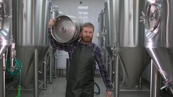 Worker Male brewer in uniform carries a keg filled with beer as he passes beer tanks video