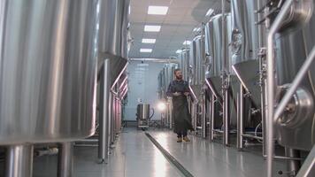 A young male brewer with a red beard walks through a craft beer factory and checks beer tanks using a tablet video