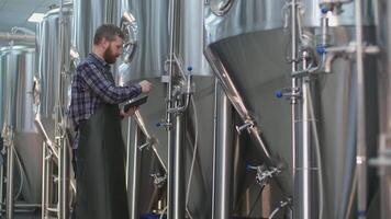 Adult Brutal Male brewer with a beard controls the readings of devices on beer tanks using a tablet. Craft beer production video