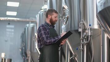 Young Brutal Male brewer with a beard stands near beer tanks and records instrument readings. Craft beer production video