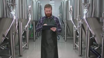 A male brewer with a beard in an apron passes by beer tanks and checks the readings using a tablet video