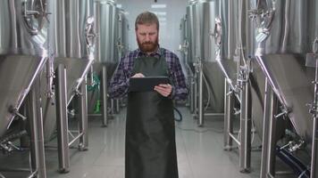 A male brewer with a beard in an apron passes by beer tanks and checks the readings using a tablet video