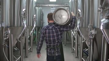 Worker A male brewer in uniform carries a keg filled with beer as he passes beer tanks. back view video