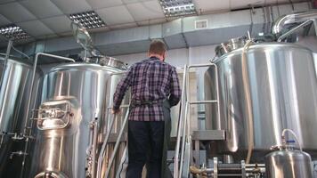 Adult Male brewer in an apron with a beard climbs the stairs to the beer tank, looks inside and records video