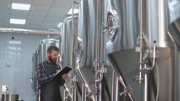 adulto maschio birraio nel un grembiule con un' barba passeggiate attraverso il birra fabbrica e record il letture su birra carri armati. produzione di mestiere birra video