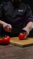 Professional chef, wearing a black uniform, throws tomatoes onto the table and then picks up one of them, deftly slicing it into thin slices with a large knife. Vertical. Close up. 4k video