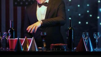 Young man in a suit is getting ready for a festive occasion and checking the table for the party. There are wine glasses, a cake, and party hats on the table. Medium shot. video