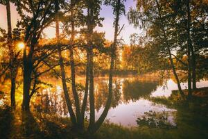 Dawn on a lake or river with a sky reflected in the water, birch trees on the shore and the sunbeams breaking through them and fog in autumn. Aesthetics of vintage film. photo