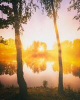 amanecer en un lago o río con un cielo reflejado en el agua, abedul arboles en el apuntalar y el rayos de sol rotura mediante ellos y niebla en otoño. estética de Clásico película. foto