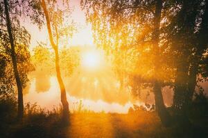 Dawn on a lake or river with a sky reflected in the water, birch trees on the shore and the sunbeams breaking through them and fog in autumn. Aesthetics of vintage film. photo