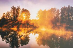 amanecer en un lago o río con un cielo reflejado en el agua, abedul arboles en el apuntalar y el rayos de sol rotura mediante ellos y niebla en otoño. estética de Clásico película. foto