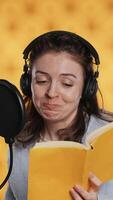 Vertical Happy narrator wearing headphones reading aloud from book into mic against yellow background. Joyous lady recording audiobook, creating engaging media content for listeners, camera A video