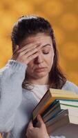 Vertical Woman with worried facial expression feeling migraine pain, carrying books for school exam, studio background. Stressed student with textbooks suffering headache from too much homework, camera A video