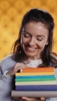 verticaal portret van gelukkig vrouw met stack van boeken in handen tonen duimen omhoog, studio achtergrond. vreugdevol boekenwurm Holding stapel van romans, gevoel vrolijk, aan het doen positief hand- gebaren, camera b video