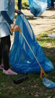 klein meisje verzamelen onzin in de omgeving van een Woud met een klauw gereedschap en vuilnis Tassen, schoonmaak omhoog de milieu door recycling plastic verspilling en afval. kind aan het leren naar beschermen ecosysteem. camera a. video