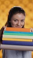 Vertical Portrait of friendly woman offering stack of textbooks useful for school exam, isolated over studio background. Merry person giving pile of books, recommending them for university assessment, camera B video