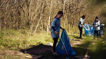 pequeño niña coleccionar basura alrededor un bosque con un garra herramienta y basura bolsas, limpieza arriba el ambiente por reciclaje el plastico residuos y basura. niño aprendizaje a proteger ecosistema. cámara b. video