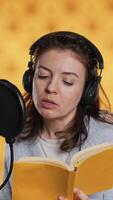 Vertical Narrator wearing headset reading aloud from book into mic against yellow background. Upbeat professional voice actor recording audiobook, creating engaging media content for listeners, camera A video