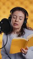 Vertical Narrator wearing headset reading aloud from book into mic against yellow background. Upbeat professional voice actor recording audiobook, creating engaging media content for listeners, camera B video