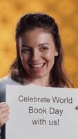 Vertical Portrait of happy woman holding placard with world book day message written on it, isolated over studio background. Geek promoting importance of reading during 23th April global event, camera A video