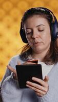 Vertical Woman holds ebook reader, listening music and purchasing books online. Person with tablet in hands buying digital novel with credit card, hearing songs in headphones, studio background, camera B video