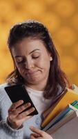 Vertical Upset woman holding stack of book, preparing for school exam using phone, studio background. Student with pile of textbooks pouting while researching for university homework using smartphone, camera A video