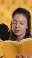 verticale influenceur tournage en train de lire livre avec téléphone intelligent sur trépied, création livre audio, studio Contexte. femme vlogging avec mobile téléphone tandis que produisant numérique enregistrement sur roman en utilisant microphone, caméra une video