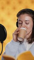 Vertical Voice actor reads book while enjoying coffee, recording audiobook using microphone, studio background. Woman using mic and headphones to produce digital recording of novel, drinking beverage, camera A video