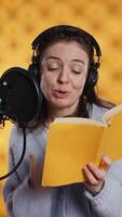 Vertical Happy narrator wearing headphones reading aloud from book into mic against yellow background. Joyous lady recording audiobook, creating engaging media content for listeners, camera B video