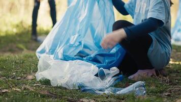 Tired little girl collecting trash and plastic bottles from the forest, helping the volunteers with litter cleanup. Child activist picking up rubbish in a garbage disposal bag. Close up. Camera A. video