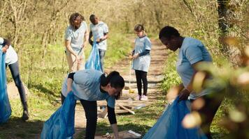 Diverse group of activists gathering to clean up a forest area, using litter cleanup tools like garbage bags and a long claw to grab the trash. People picking rubbish and recycling. Camera A. video