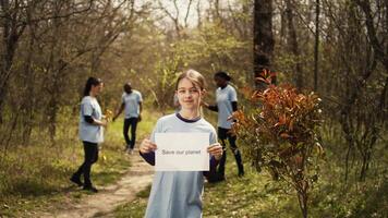 retrato de dulce niña con salvar nuestra planeta póster en contra contaminación y ilegal dumping, trabajar como voluntario a restaurar y preservar naturaleza en el bosque. pequeño niño muestra conciencia signo. cámara b. video