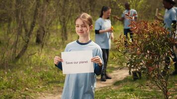 Porträt von Süss Mädchen mit speichern unser Planet Poster gegen Verschmutzung und illegal Schluss machen, Freiwilligenarbeit zu wiederherstellen und erhalten Natur im das Wald. wenig Kind zeigt an Bewusstsein unterzeichnen. Kamera a. video