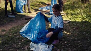 cansado pequeno menina colecionar Lixo e plástico garrafas a partir de a floresta, ajudando a voluntários com lixo limpar. criança ativista colheita acima lixo dentro uma lixo disposição bolsa. fechar acima. Câmera b. video