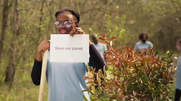 Porträt von afrikanisch amerikanisch Aktivist halten Poster mit speichern unser Planet Nachricht, Verbreitung Umwelt Pflege und Bewusstsein. jung Frau posieren mit Banner zu Kampf ökologisch Gerechtigkeit. Kamera a. video