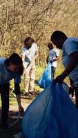 Diverse group of activists gathering to clean up a forest area, using litter cleanup tools like garbage bags and a long claw to grab the trash. People picking rubbish and recycling. Camera B. video