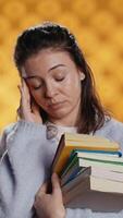 Vertical Woman with worried facial expression feeling migraine pain, carrying books for school exam, studio background. Stressed student with textbooks suffering headache from too much homework, camera B video