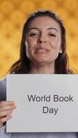 Vertical Smiling book club member holding placard with world book day message, isolated over studio background. Radiant bookworm recording promoting literacy importance, camera B video