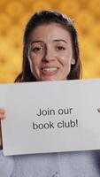 Vertical Cheerful book club president holds message urging people to join them, talking about importance of lecture, studio background. Smiling woman inviting bookworms to enlist in her association, camera B video