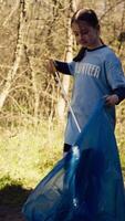 Young child using tongs tool to grab and pick up trash from the woods, collecting rubbish from a natural habitat. Little girl growing up with environmental care and recycling plastic waste. Camera B. video