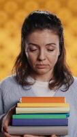 verticaal portret van glimlachen vrouw Holding stack van boeken, aan het doen begroeting hand- gebaar, gevoel optimistisch. vrolijk persoon met stapel van romans verhogen arm naar begroeten iemand, studio achtergrond, camera b video