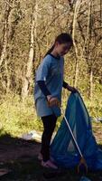 pequeno menina colecionar lixo por aí uma floresta com uma garra ferramenta e lixo bolsas, limpeza acima a meio Ambiente de reciclando plástico desperdício e lixo. criança Aprendendo para proteger ecossistema. Câmera b. video