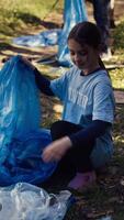 moe weinig meisje verzamelen uitschot en plastic flessen van de Woud, helpen de vrijwilligers met afval schoonmaken. kind activist plukken omhoog onzin in een vuilnis verwijdering tas. dichtbij omhoog. camera b. video