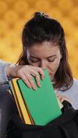 vertical retrato do mulher vasculhando através mochila, levando livro fora, isolado sobre estúdio fundo. aluna removendo livro didático usava para educacional finalidades a partir de escola mochila, Câmera b video