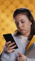 Vertical Upset woman holding stack of book, preparing for school exam using phone, studio background. Student with pile of textbooks pouting while researching for university homework using smartphone, camera B video