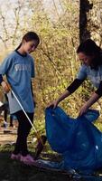 Mom and child collecting rubbish from the forest area using a long claw tool and garbage disposal bags. Little girl picking up trash and plastic waste with her mother, ecological justice. Camera B. video