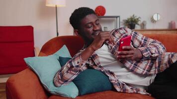 Cheerful young man lying on sofa, using mobile phone share messages on social media application video