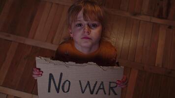 Scared homeless toddler girl sitting holding inscription No War, hiding from bombing attack at home video