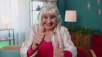 Overjoyed senior woman in wireless headphones dancing, singing on cozy couch in living room at home video